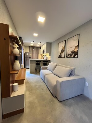 Un salon moderne avec un canapé, une étagère en bois et une cuisine en arrière-plan.