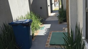 Een smal pad, begrensd door gebouwen, met een blauwe prullenbak en groene planten.