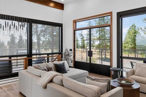 A modern living room with a sofa, a coffee table, and large windows offering a view of trees and a patio. - Panoramic River Views, Luxury Kitchen & Fireplace (Chelan)