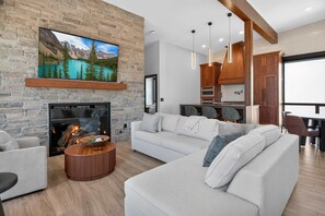A modern living room with a stone fireplace, a large flat-screen TV, a white sofa, and a dining area with wooden cabinets. - Panoramic River Views, Luxury Kitchen & Fireplace (Chelan)