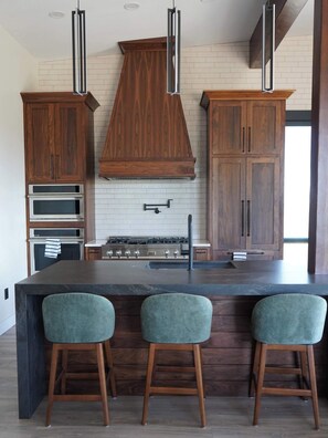A modern kitchen with wooden cabinets, a central island with bar stools, a built-in oven, and a stainless steel range.