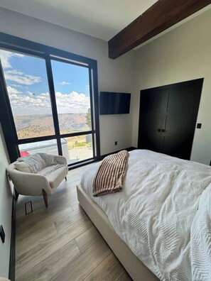 A bedroom with a large bed, a sofa, a TV, and a view of a landscape through a large window.