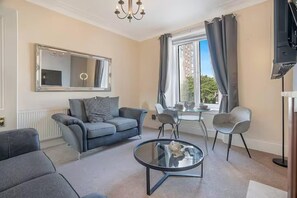 Ein Wohnzimmer mit einer Couch, zwei Sesseln, einem Couchtisch, einem Fernseher und einem großen Fenster mit Vorhängen.