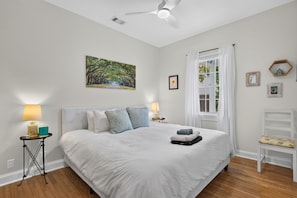 A bedroom with a large bed, a ceiling fan, a window with curtains, and a painting on the wall. - Newly Refreshed | King Bed | Walk to Forsyth (Savannah)
