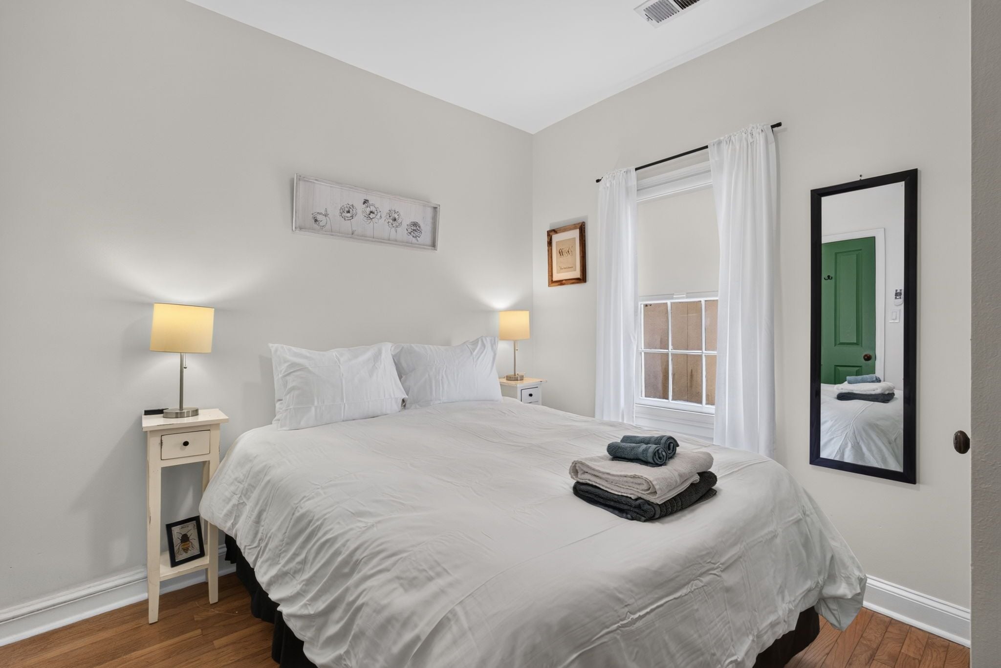 Un dormitorio con una cama grande, mesitas de noche, una ventana con cortinas, un espejo y una pared con un cuadro.