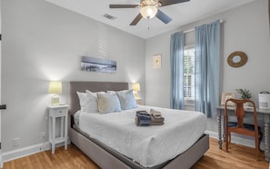 Une chambre à coucher comprenant un lit, des lampes de chevet, un ventilateur de plafond, une fenêtre avec des rideaux et une petite table avec une chaise.