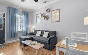 Un salon avec un canapé gris, une table basse en bois, une petite table d’appoint avec une lampe et une chaise blanche. Des tableaux encadrés ornent le mur, et une fenêtre est agrémentée de rideaux bleus.
