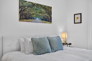 A bedroom with a bed, pillows, a nightstand with a lamp, and two framed pictures on the wall. - Historic Cottage | Walk to Forsyth | King + Queen (Savannah)