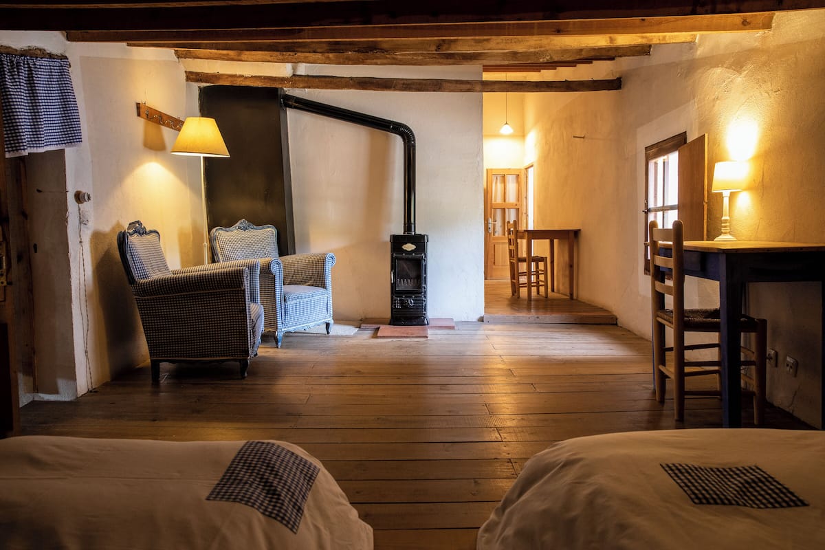 A cozy room with wooden floors, two beds, a chair, a table, and a fireplace.