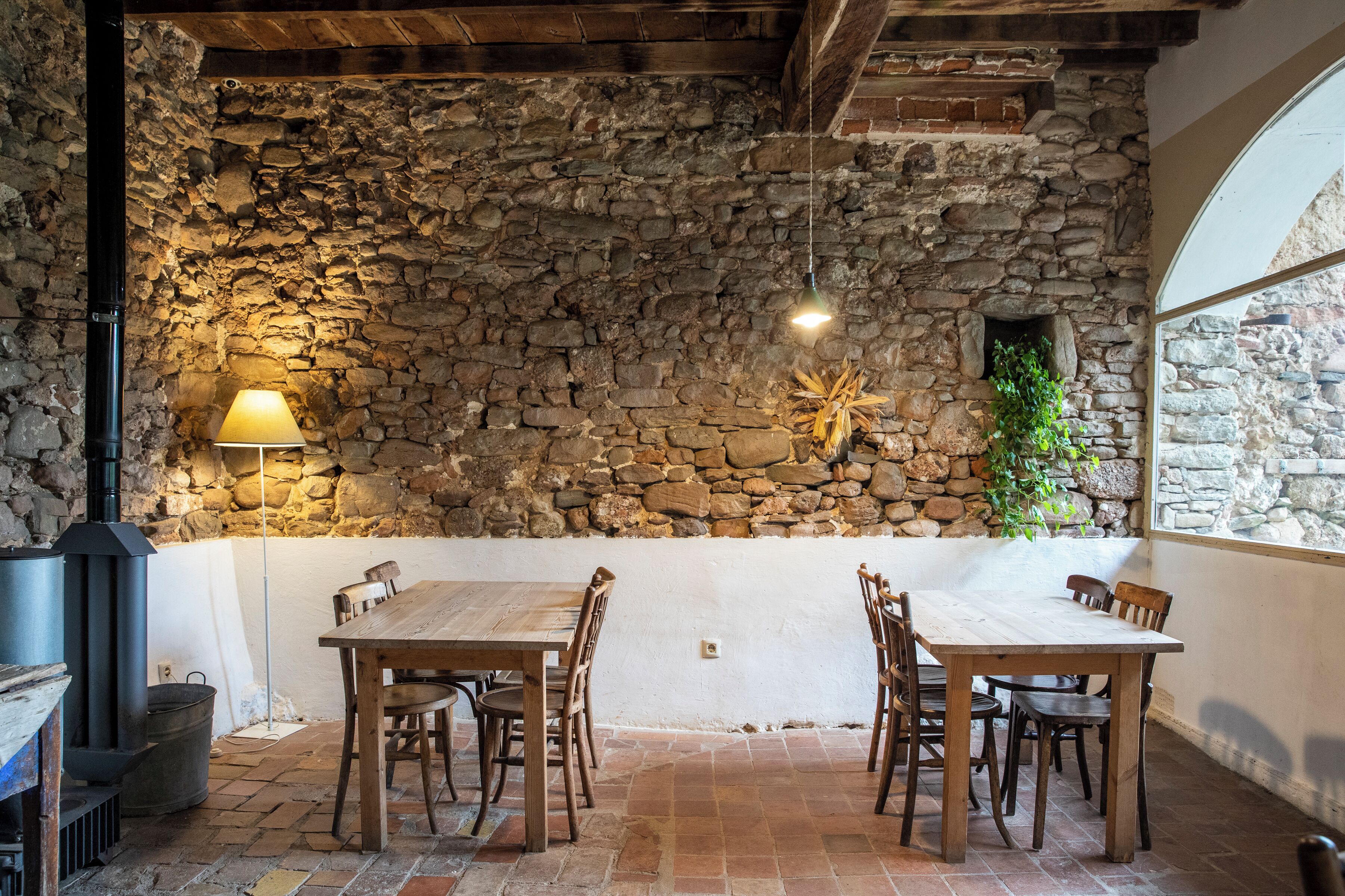 A rustic stone wall interior with a wooden table and chairs, a stone fireplace, and a window providing natural light.