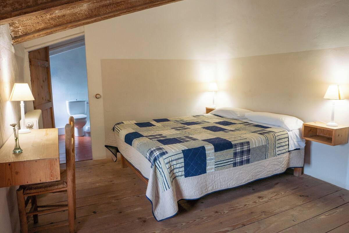A bedroom with a wooden floor, a bed with a quilt, a wooden chair, and two bedside lamps.