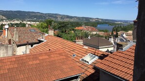 On aperçoit des toits recouverts de tuiles en terre cuite, une étendue d’eau et des collines au loin.