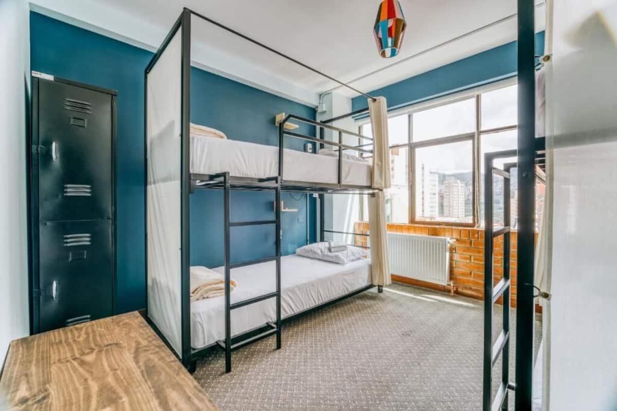 A dormitory room with bunk beds, a wardrobe, a radiator, and a window with a city view.