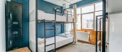 A dormitory room with bunk beds, a wardrobe, a radiator, and a window with a city view.