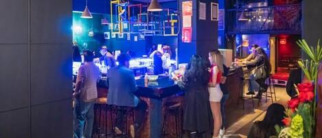 Zona de bar con taburetes, una barra y gente interactuando.