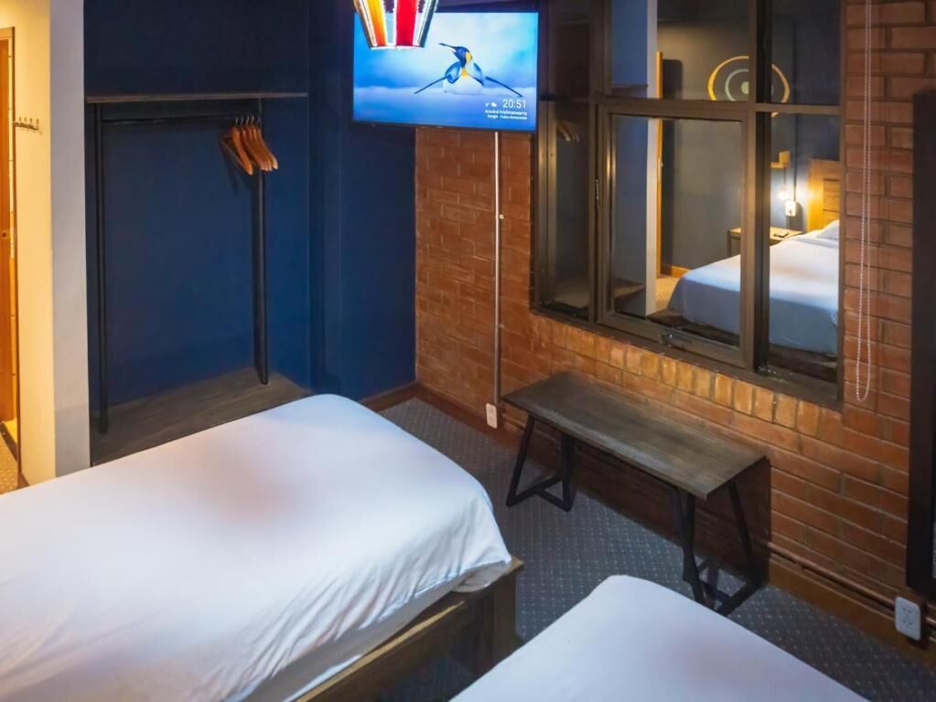 Ein Hotelzimmer mit zwei Betten, einem an der Wand montierten Fernseher, einer Bank und einem Fenster mit Blick auf den Raum.