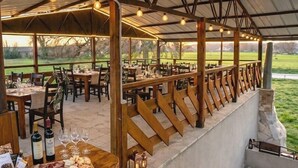An outdoor dining area with wooden tables and chairs, a stone wall, and a view of a green field.