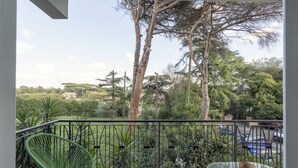 En balkon med to stole, et glasbord og udsigt til træer samt en bygning i det fjerne.