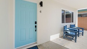 Eine blaue Tür mit der Zahl 4 darüber, eine kleine Außenbereich-Sitzgelegenheit mit zwei Stühlen und einem Tisch sowie ein Fenster mit Blick auf eine Ziegelwand.