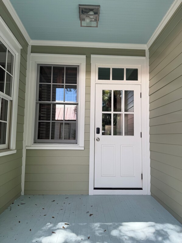 A porch with a white door and two windows, a light blue ceiling, and a wooden floor with scattered leaves.