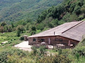A rustic wooden building with a sloped roof, surrounded by lush greenery and hills. - La Ferme du Maquis (Antisanti)