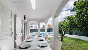 An outdoor dining area with a white table set for six, surrounded by white chairs, under a covered structure with a view of a swimming pool and greenery. - Villa Martins with Private Pool, Wi-Fi, and Air Conditioning (Albufeira)