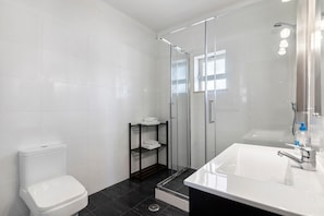A bathroom with a white toilet, a glass-enclosed shower, a white sink with a mirror, and a shelf with towels.