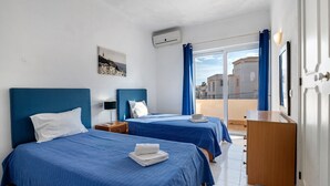 A hotel room with two beds, a small desk, a window with curtains, and a wall-mounted air conditioner. - Villa Martins with Private Pool, Wi-Fi, and Air Conditioning (Albufeira)