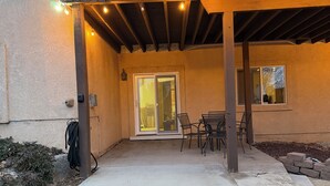 Een terras buiten met een betonnen vloer, een pergola met lichtslingers, een schuifdeur en een raam.