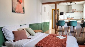 A room with a bed, a green upholstered headboard, a dining area with two bar stools, and a kitchen visible through an open doorway. - L'Echappée Urbaine Rouennaise - Hyper downtown - Rouen Left Bank Station (Rouen)