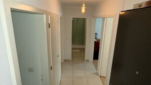 En smal hall med vita väggar och ett kaklat golv, som leder till ett badrum med dusch och toalett.