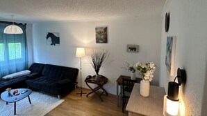 Ett vardagsrum med en mörk soffa, ett litet runt bord, en lampa och en vas med blommor.