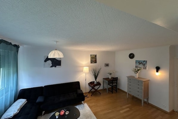 A living room with a black sofa, a small round table, a lamp, a side table with a vase, and a dresser.