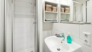Ett modernt badrum med vita kakelplattor, en dusch med glasväggar, en vit tvättställ med tvålpump och hyllor med korgar för förvaring.