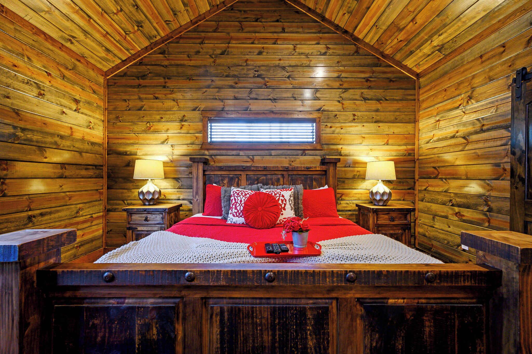 A log cabin bedroom with a wooden ceiling, a bed with red bedding, two bedside lamps, and a window with blinds.