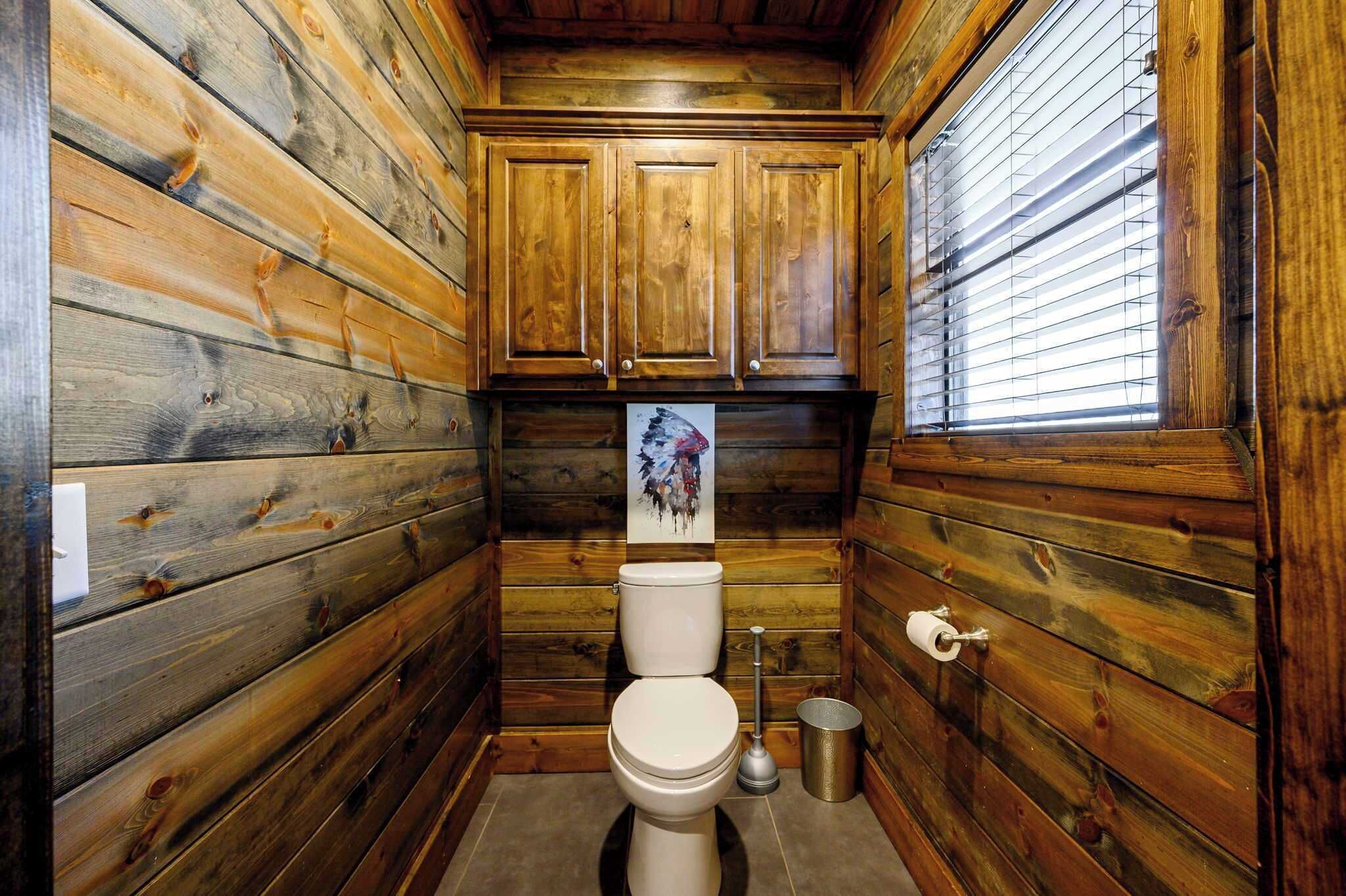 A wooden cabin-style bathroom with a toilet, a trash can, and a window with blinds.