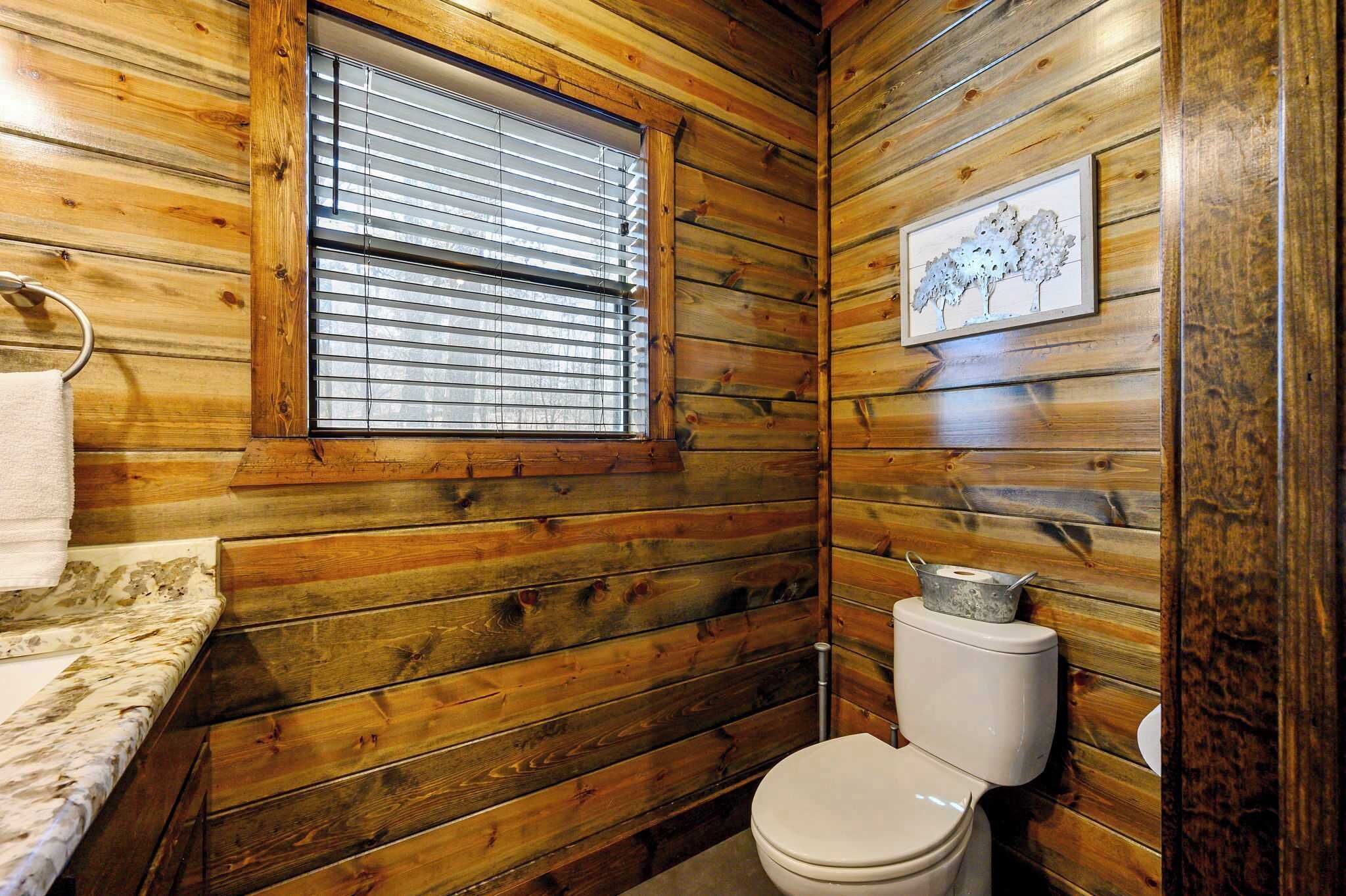 Un baño con paneles de madera, un inodoro, una ventana con persianas y un cuadro en la pared.