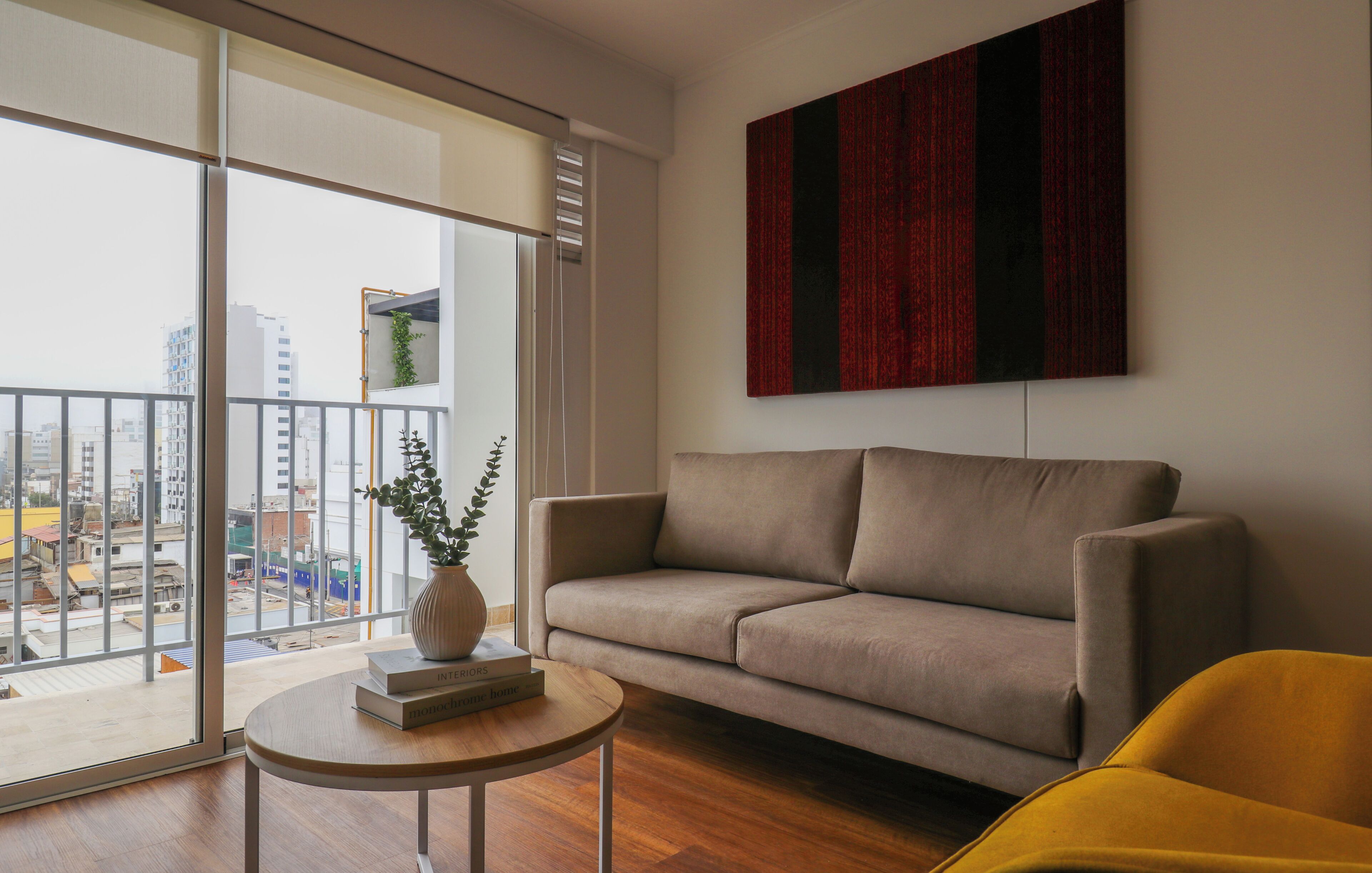Una sala de estar moderna con un sofá, un sillón amarillo, una mesa redonda de madera y un ventanal con vistas a edificios.