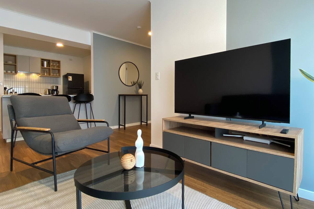 Uma sala de estar moderna com televisão de tela plana, um sofá cinza, uma mesa de centro redonda de vidro e uma cozinha ao fundo.