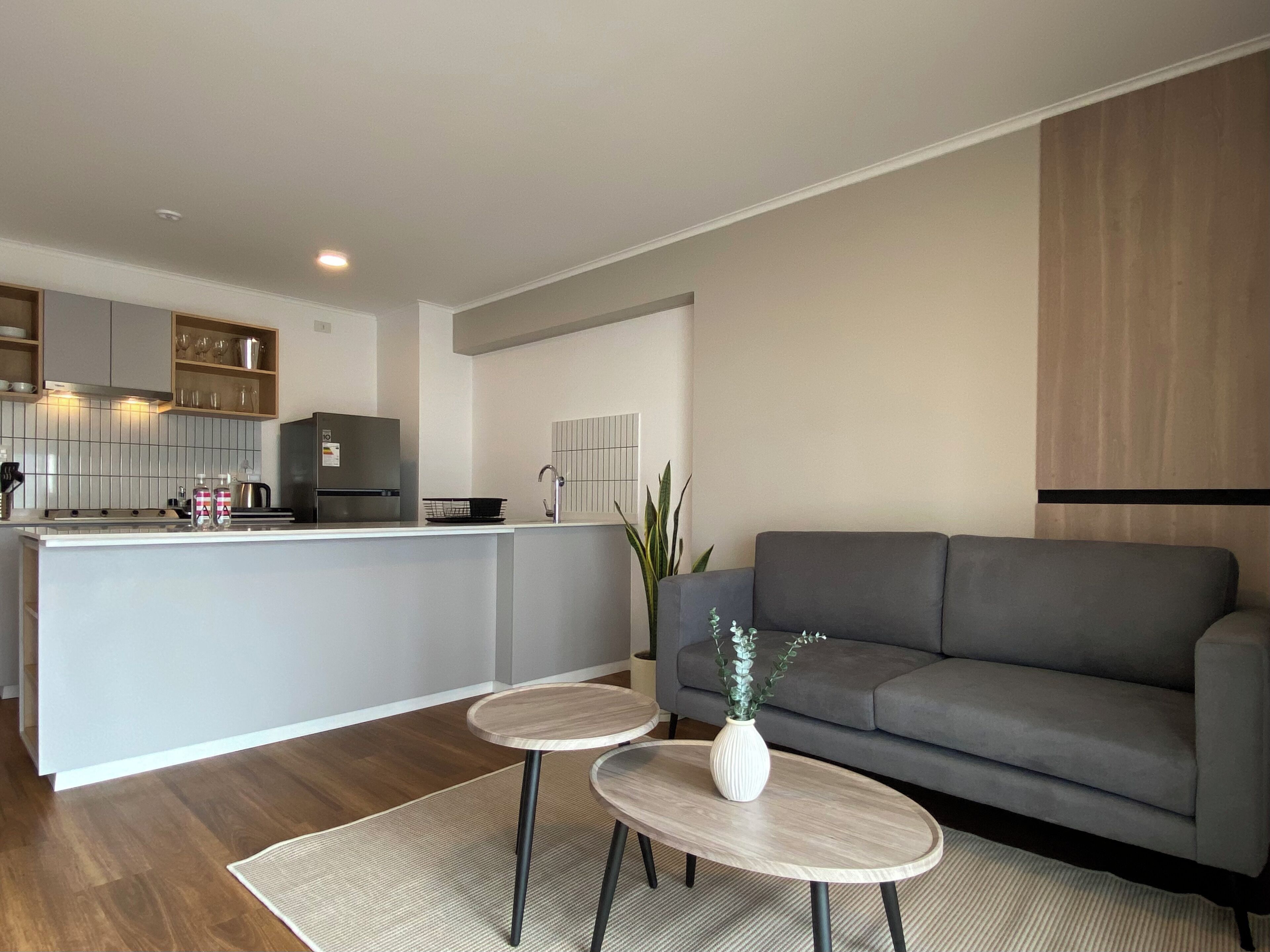 Una cocina moderna con armarios blancos, una isla gris y un sofá gris en la sala de estar. Hay dos mesas redondas con jarrones sobre ellas.