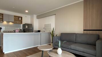 Una cocina moderna con armarios blancos, una isla gris y un sofá gris en la sala de estar. Hay dos mesas redondas con jarrones sobre ellas.