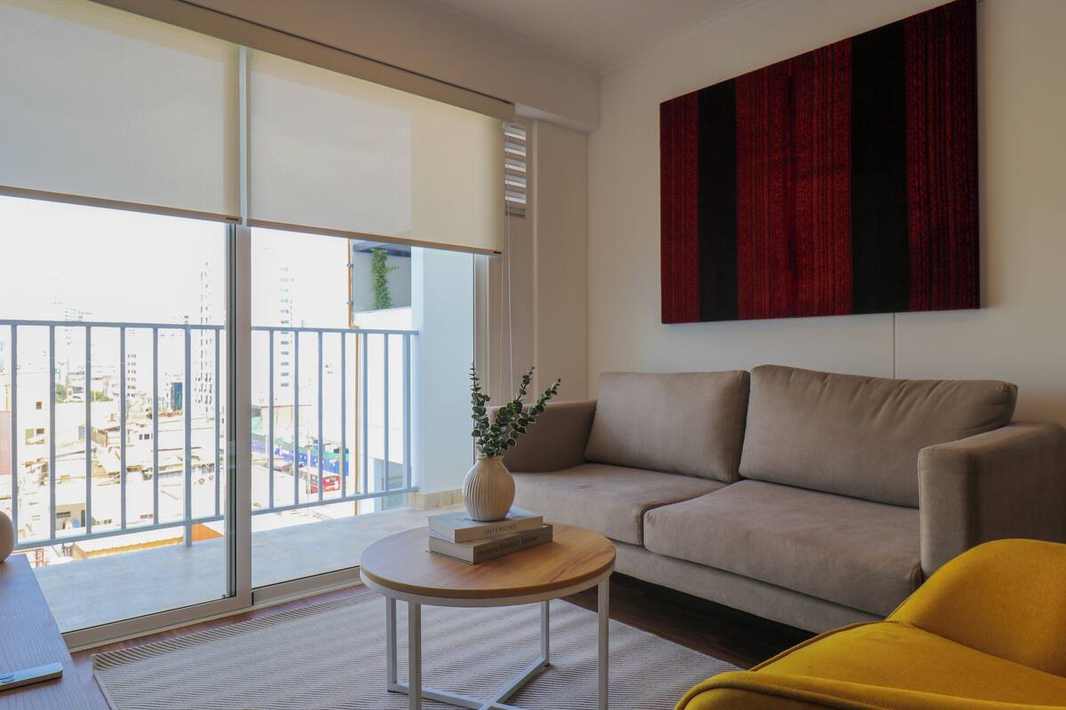 Uma sala de estar moderna com um sofá, uma poltrona amarela, uma mesinha redonda com um vaso e livros, e uma janela grande com vista para edifícios.