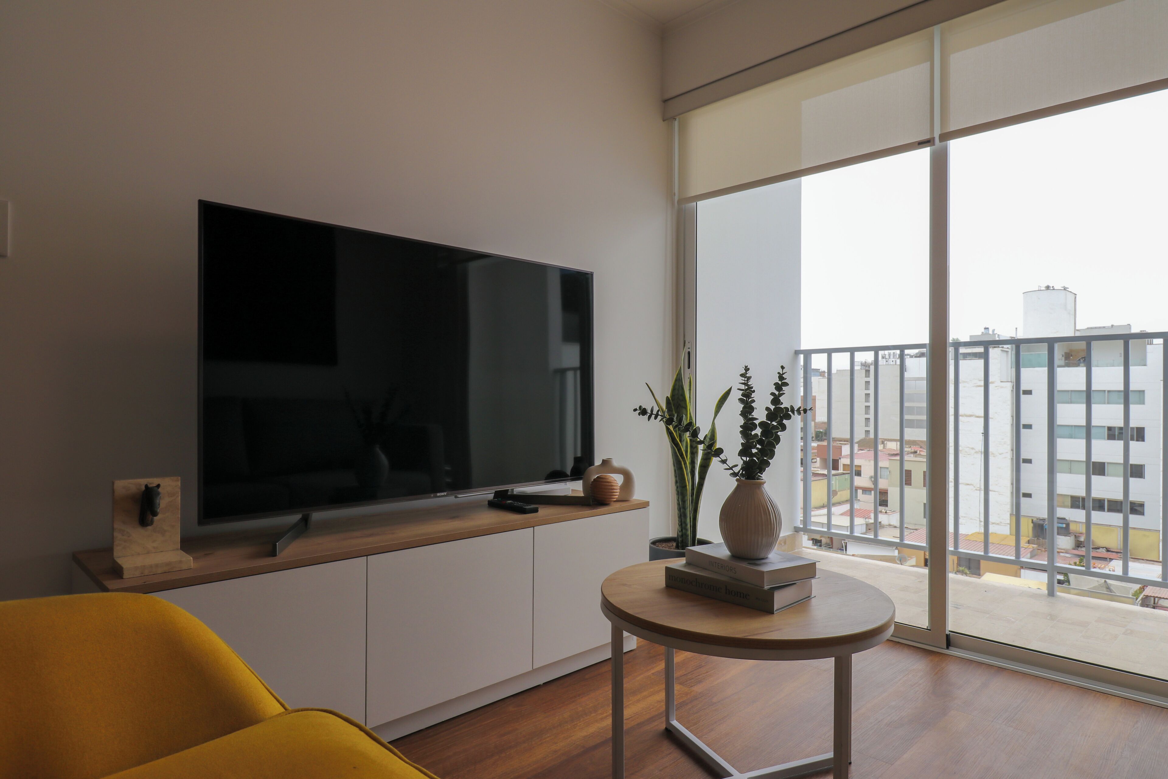 A modern living room with a flat-screen TV, a yellow sofa, a small round table with books, and a large window with a city view.
