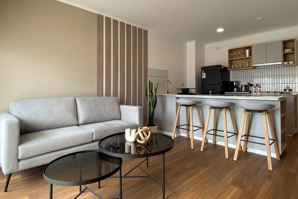 Uma sala de estar moderna com um sofá cinzento, uma mesa de centro redonda de vidro e banquetas de madeira. Uma cozinha com uma geladeira preta e armários brancos é visível ao fundo.