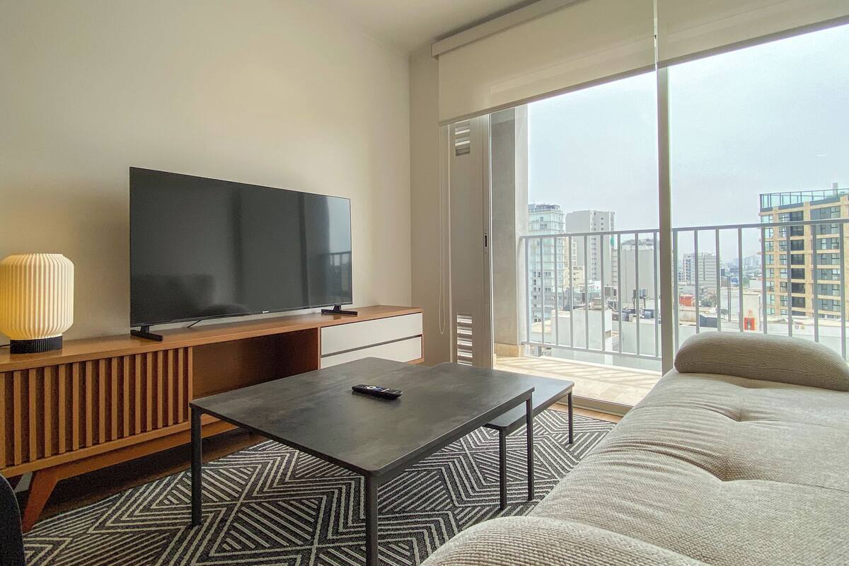 Uma sala de estar moderna com uma televisão de tela plana, um sofá, uma mesa de centro e uma varanda com vista para a cidade.