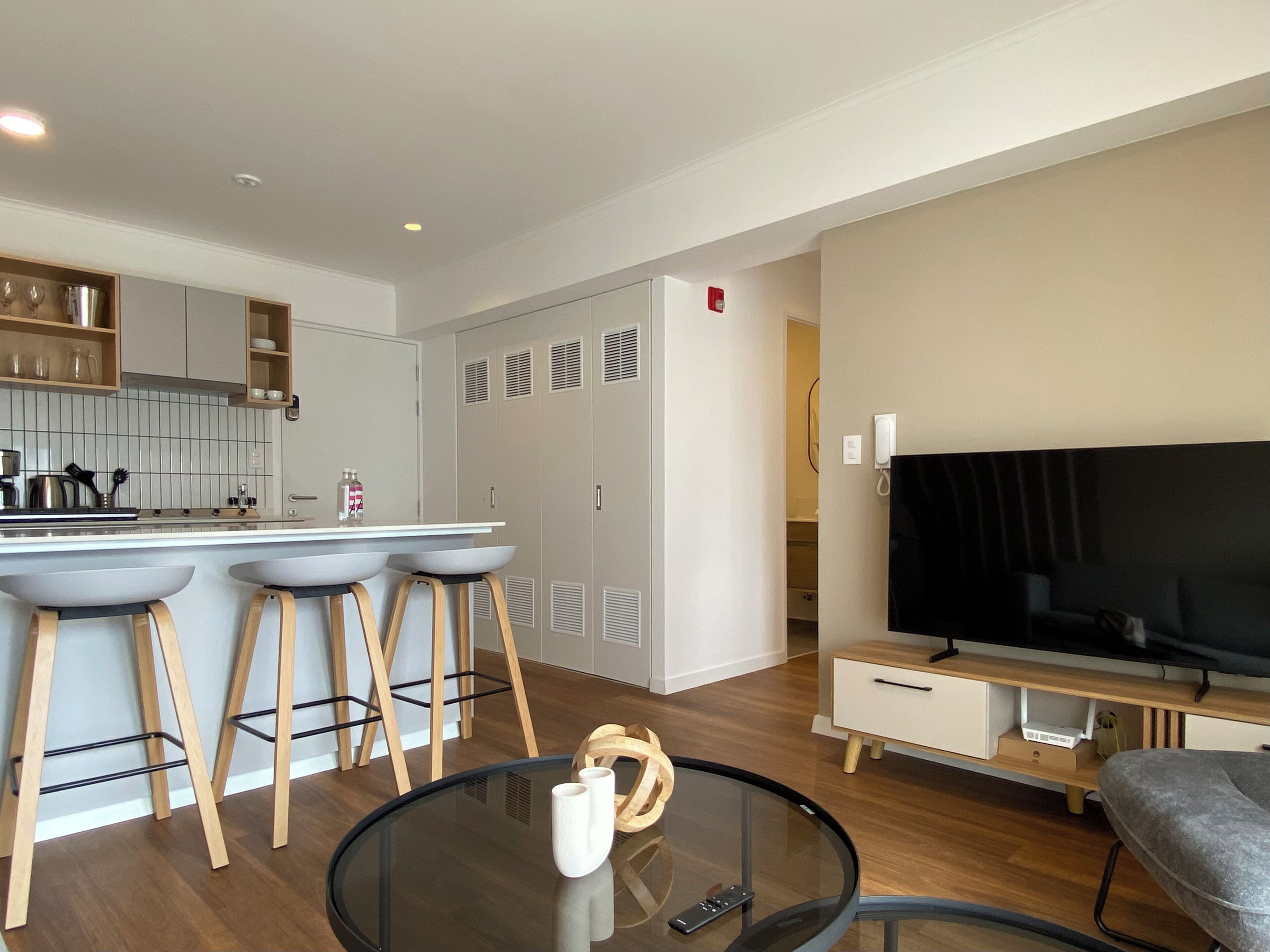 A modern apartment with a kitchen, dining area, and living room. It includes a kitchen with white cabinets, a dining table with bar stools, a flat-screen TV, and a glass coffee table.