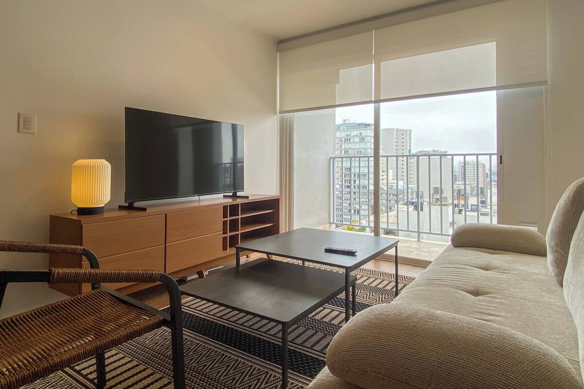 Uma sala de estar moderna com televisão de tela plana, sofá, mesa de centro e varanda com vista para a cidade.