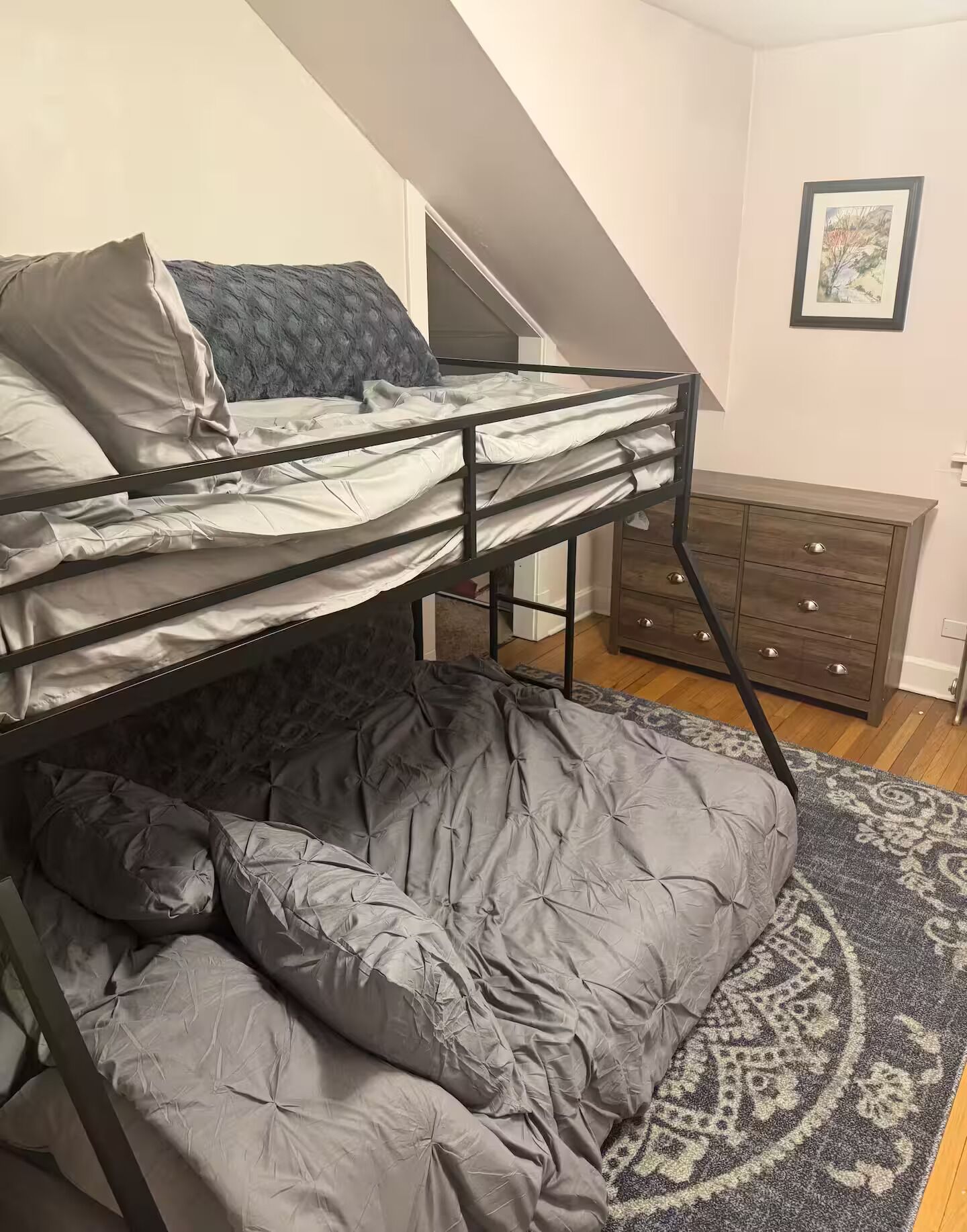 Un lit superposé avec une literie grise, un tapis à motifs et une commode en bois dans une pièce dont le plafond est en pente.