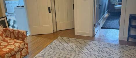 A living room with a patterned rug, a sofa, and a chair. There is a door leading to a hallway and a kitchen.