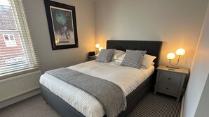 Een keurig opgemaakt slaapkamer met een bed, twee nachtkastjes met lampen, een ingelijst poster aan de muur en een raam met jaloezieën.